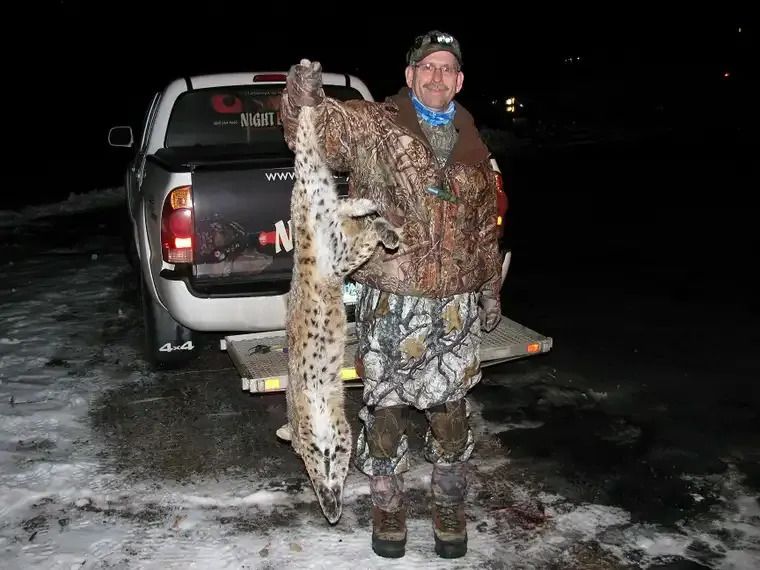Hunter holding a large spotted wild cat beside a pickup truck on icy ground at night