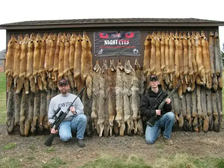 Two men kneel before a wall of hanging animal pelts and taxidermy, holding rifles in an outdoor display.