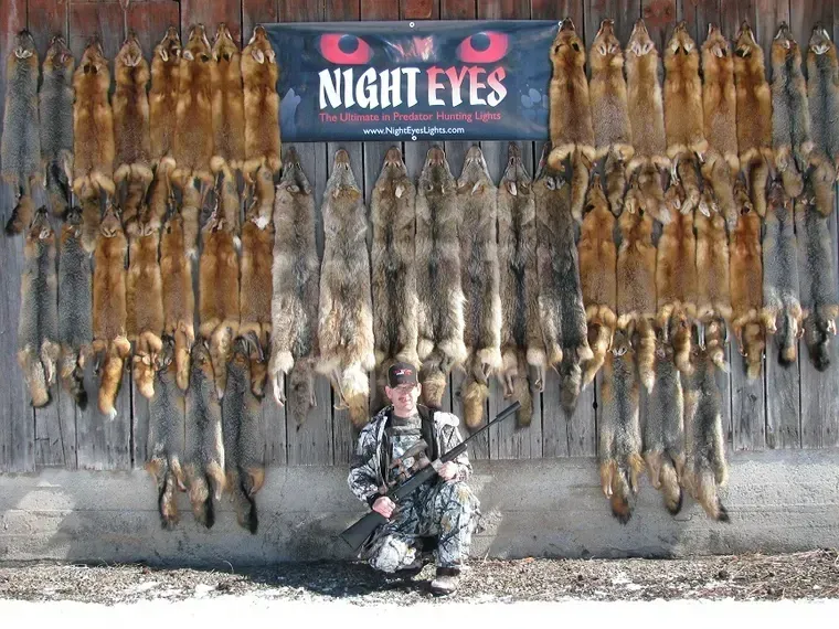 Camouflaged hunter seated beneath a wall of hanging fur pelts and a “NIGHT EYES” sign