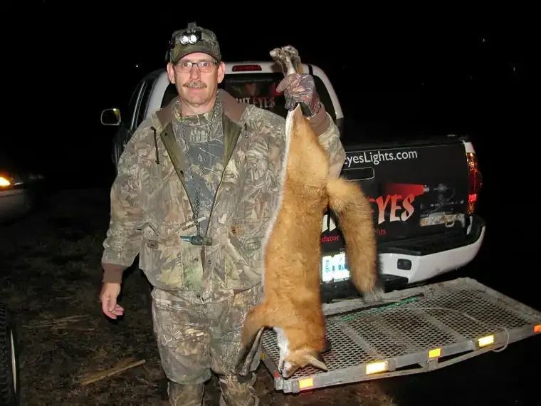 Man in camouflage holding a large fox beside a pickup truck at night