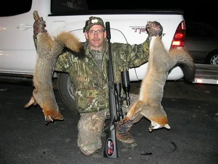 Hunter kneels at night, holding two dead foxes beside a truck, with a rifle leaning in front.