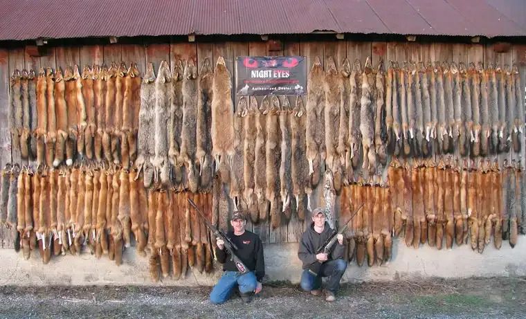 Two people kneel before rows of hanging smoked fish outside a rustic building.
