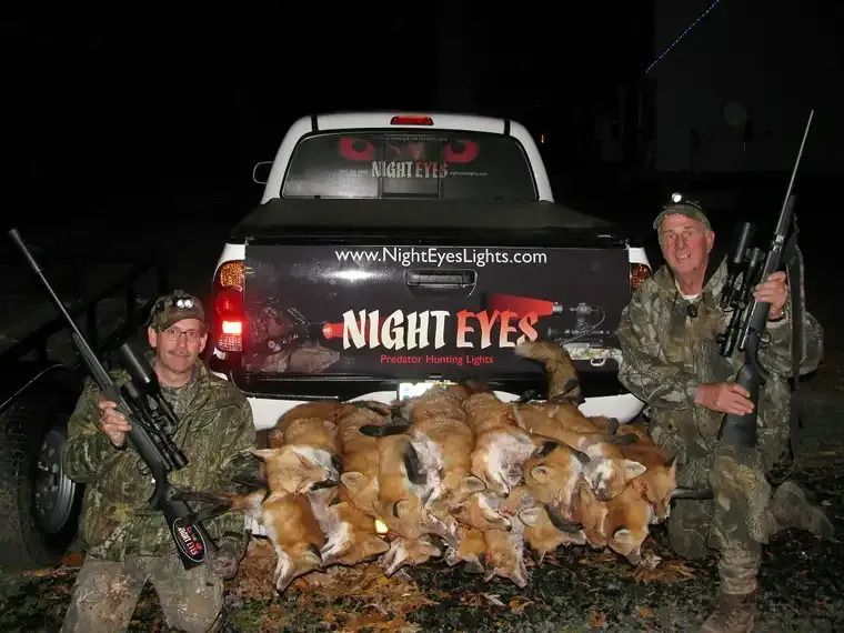 Two hunters kneel beside a pile of foxes in front of a truck marked “Night Eyes” at night