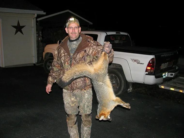 Man in camouflage holding a large fox by the tail beside a pickup truck at night