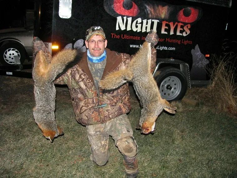 Man in camouflage holding two large fox pelts in front of a Night Eyes truck at night