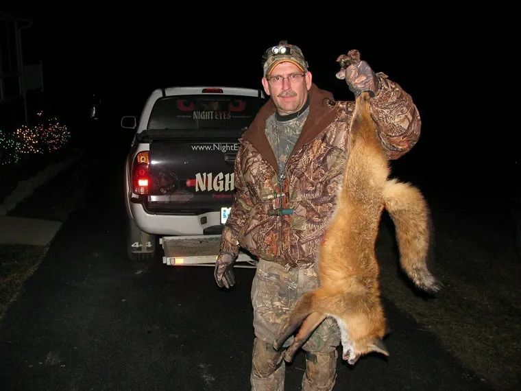 Hunter holding a dead fox by the tail beside a pickup truck at night