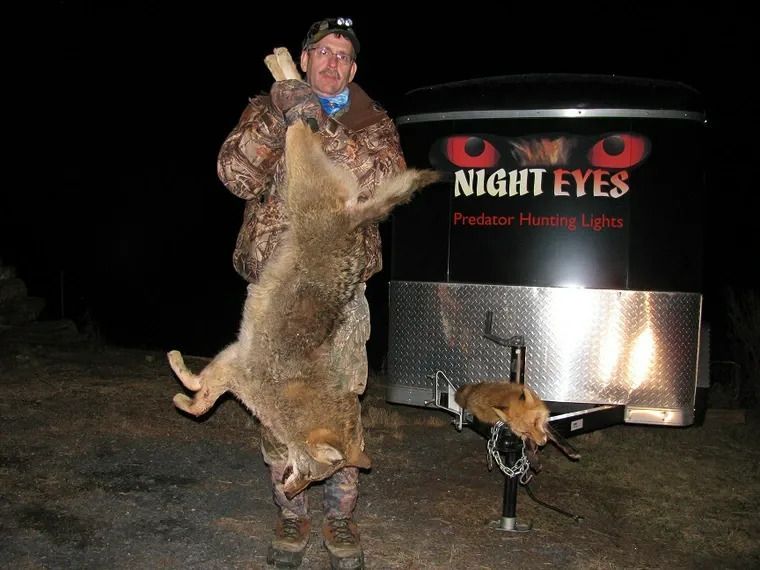 Hunter posing with a large coyote beside a lit Night Eyes trailer at night