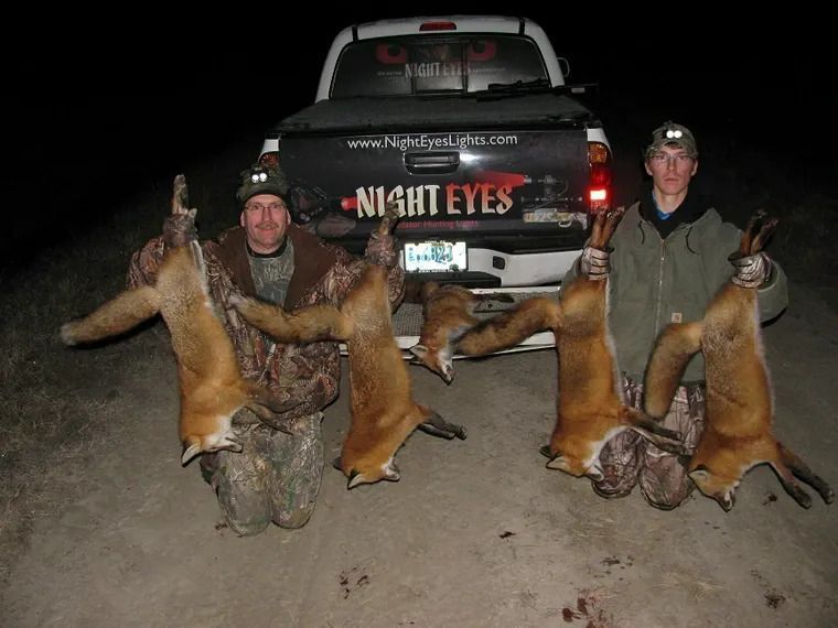 Three hunters posing with two deer carcasses in front of a truck at night