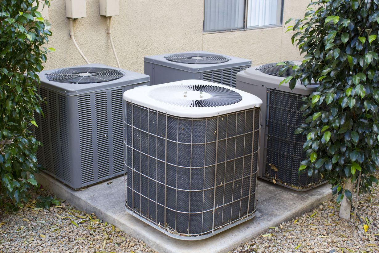 Three grey HVAC air conditioning units sitting on a concrete pad outdoors, flanked by green bushes.