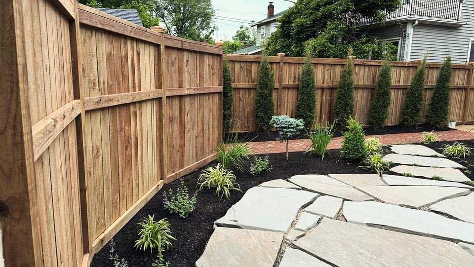 A wooden fence surrounds a stone patio in a backyard.
