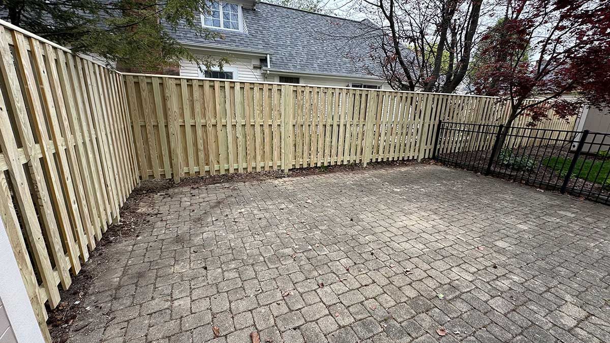 A wooden fence surrounds a brick patio in a backyard.