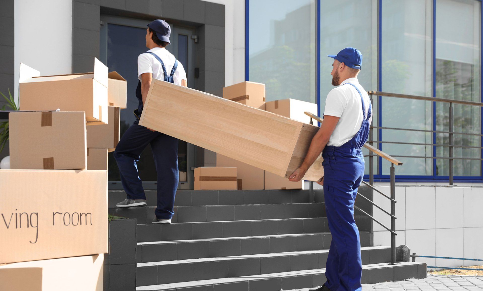 Two movers carry a long box up steps beside a moving truck loaded with cardboard boxes.