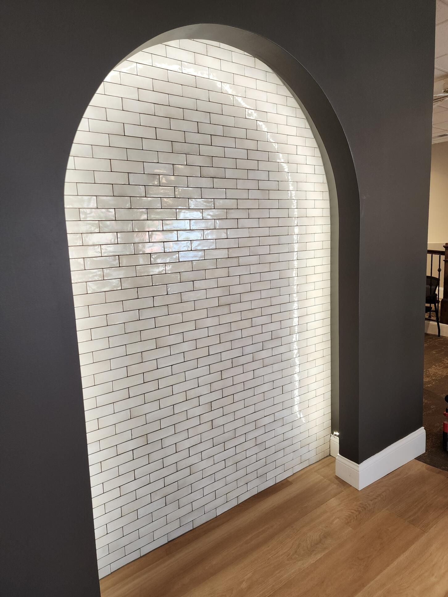 Arched recessed wall with white brick tiles, illuminated, flanked by dark gray walls, and wooden floor.
