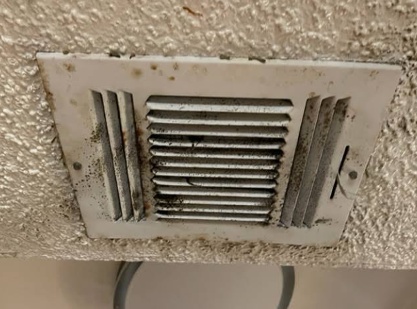 A dirty, white air vent with dark debris covering the surface and slats, mounted in a textured ceiling.