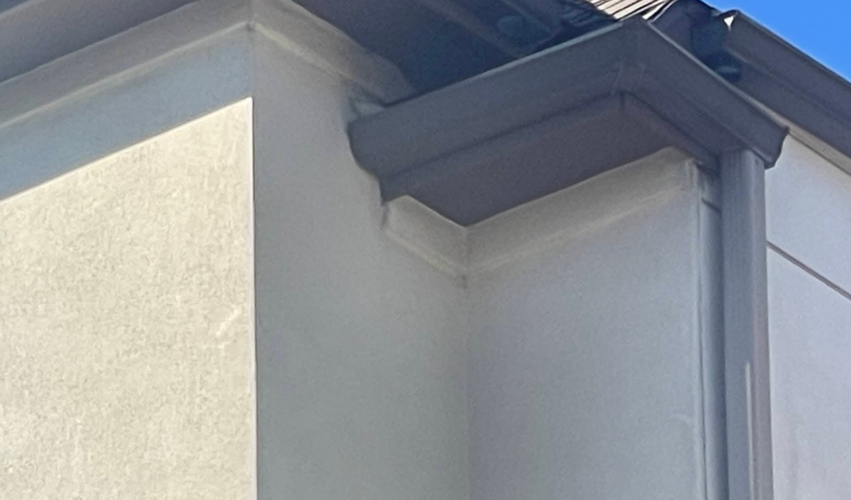 Corner of a stucco building with dark trim, gutters, and a portion of a roof.