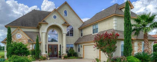 Large, elegant house with beige stucco, arched entry, brown roof, and lush landscaping against a blue sky.