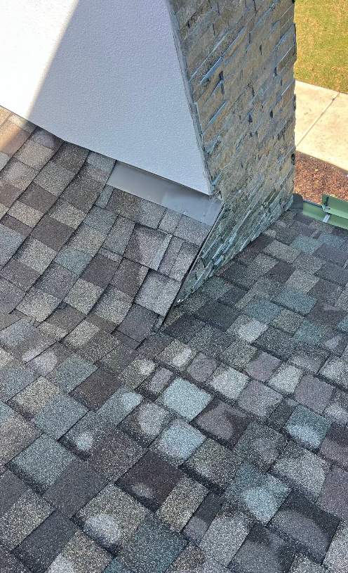 Roof shingles meeting chimney. Gray and blue roof, white house siding, stone chimney.