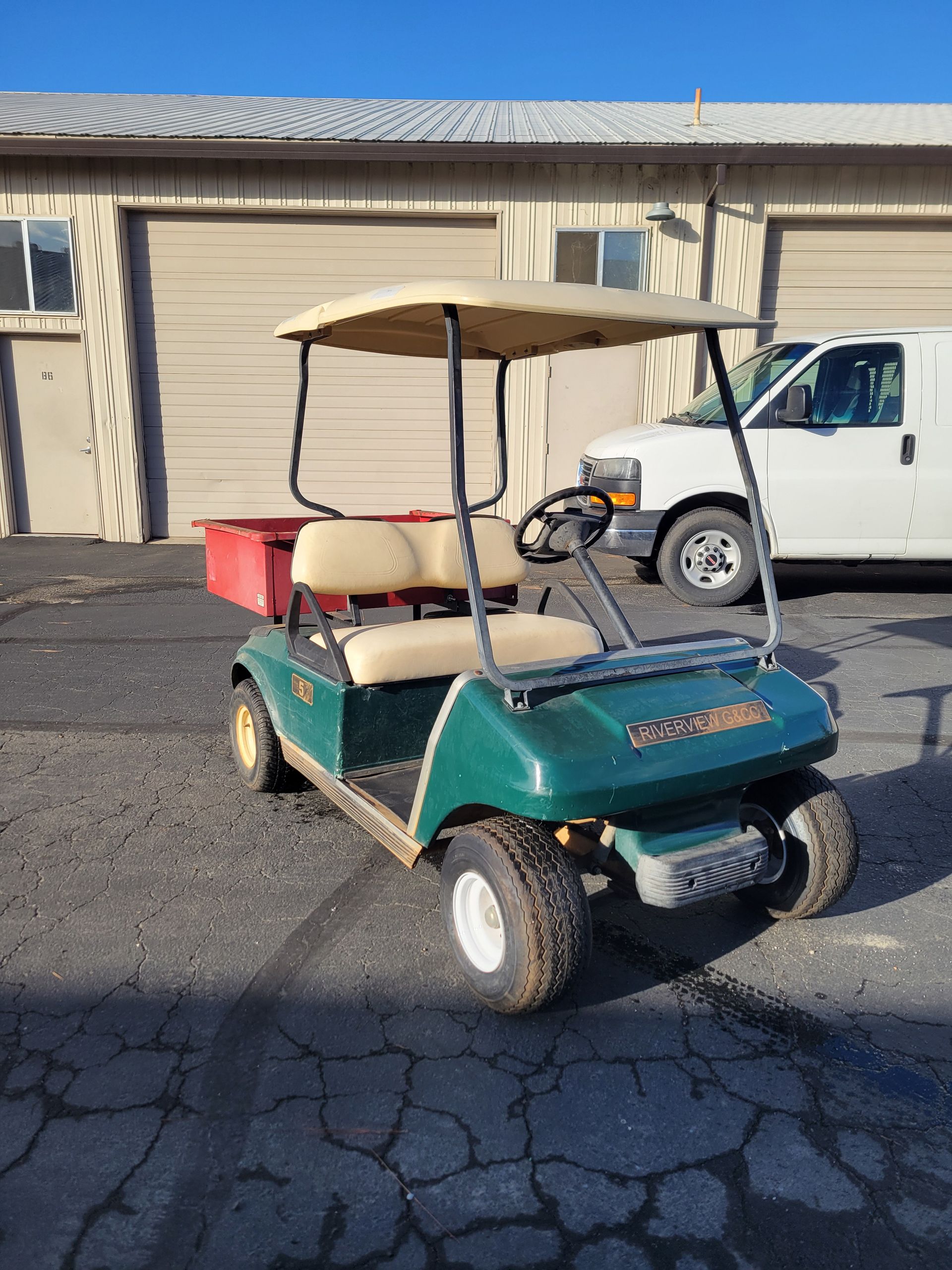 Green golf cart with tan roof, red utility box, parked in front of a building and white van.