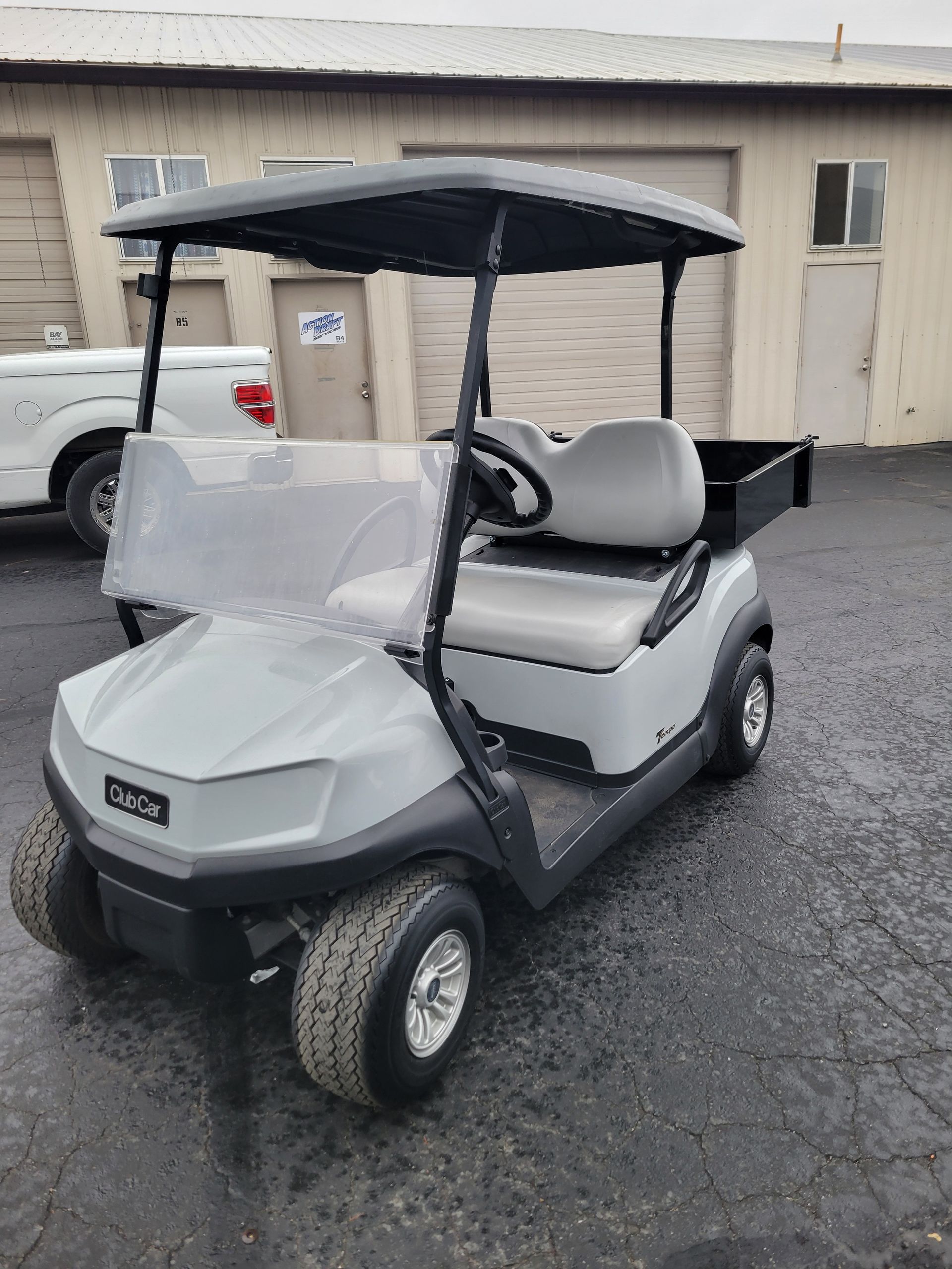 Light gray golf cart with a black canopy and cargo bed, parked outside a building.