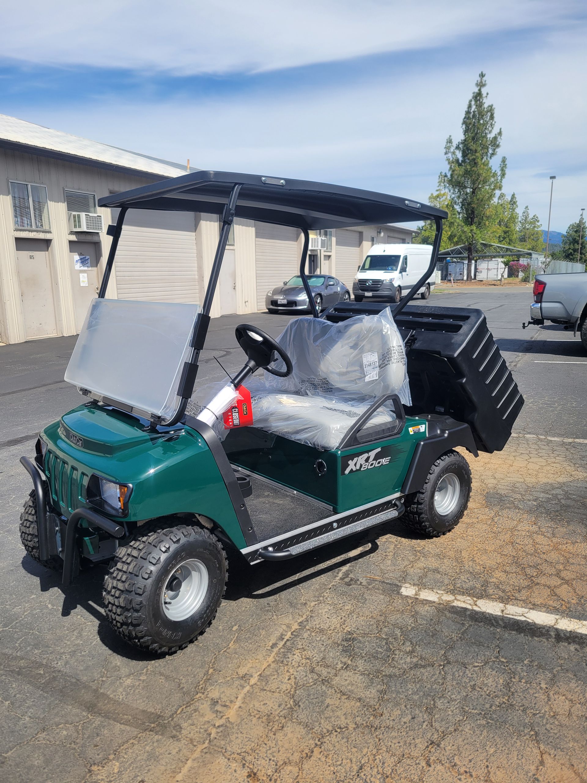 Green golf cart parked outdoors in a lot, covered in plastic with a white windshield cover.