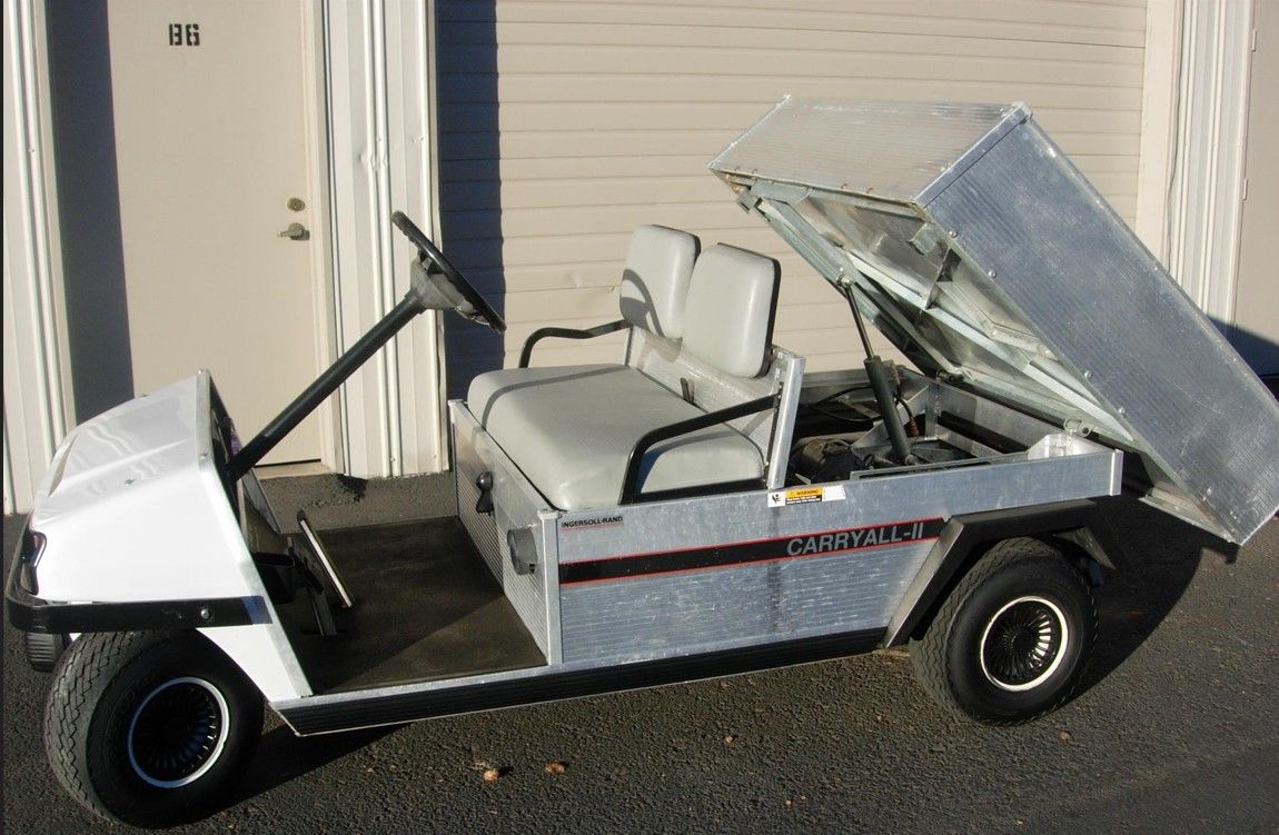 White and silver golf cart with a raised dump bed; parked outside a building.