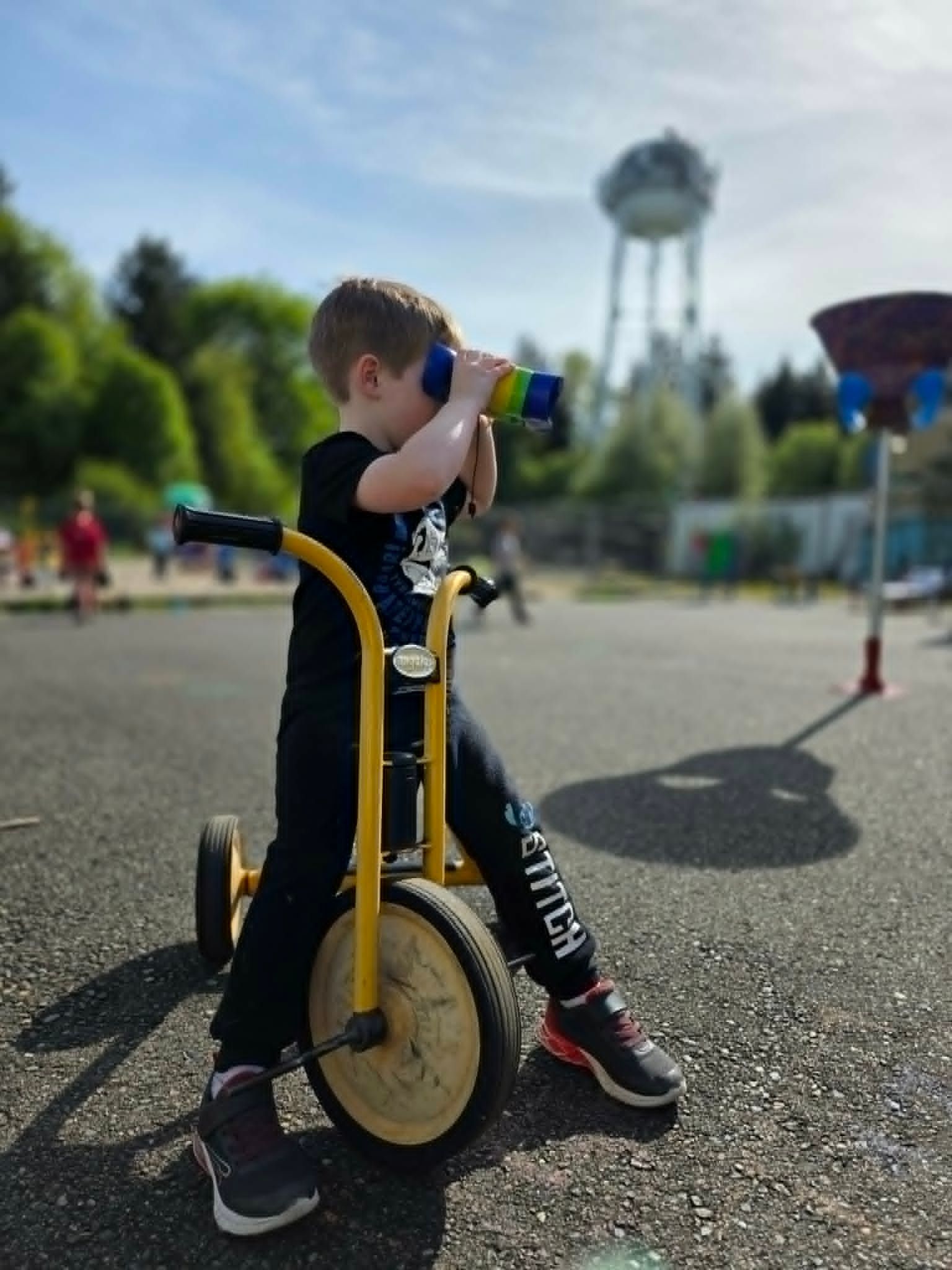 Child drinking from a blue bottle while sitting on a yellow scooter in a park