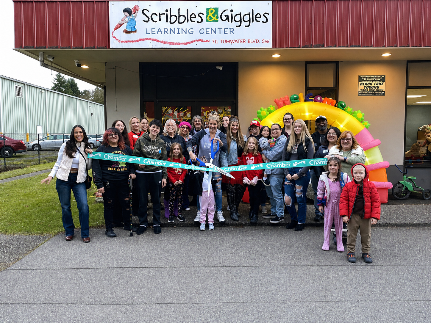 Group outside Scribbles & Giggles Learning Center, holding a ribbon at a grand opening, with a colorful balloon arch.