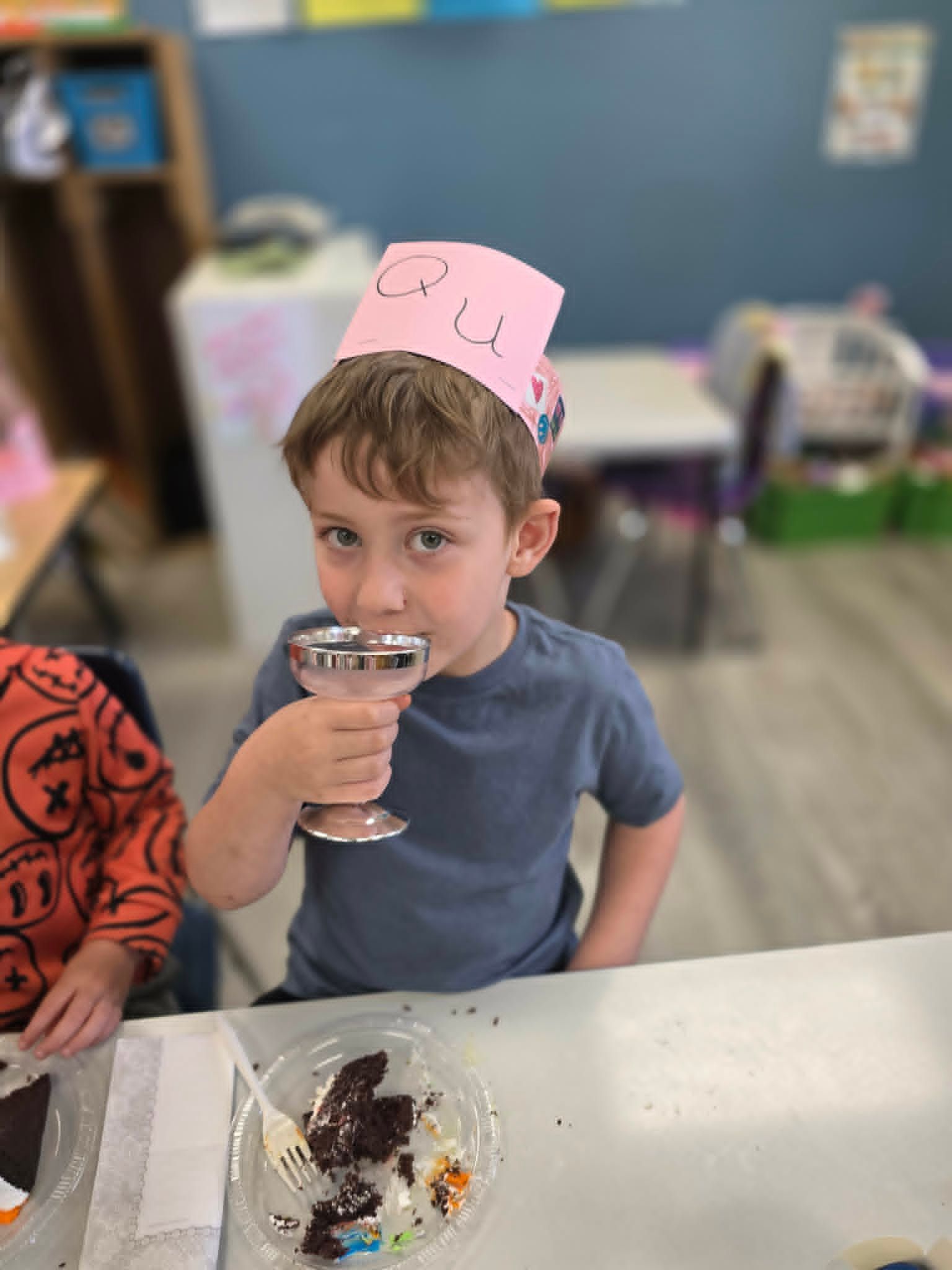 Child wearing a pink paper crown sips from a cup at a table with dessert in a classroom.