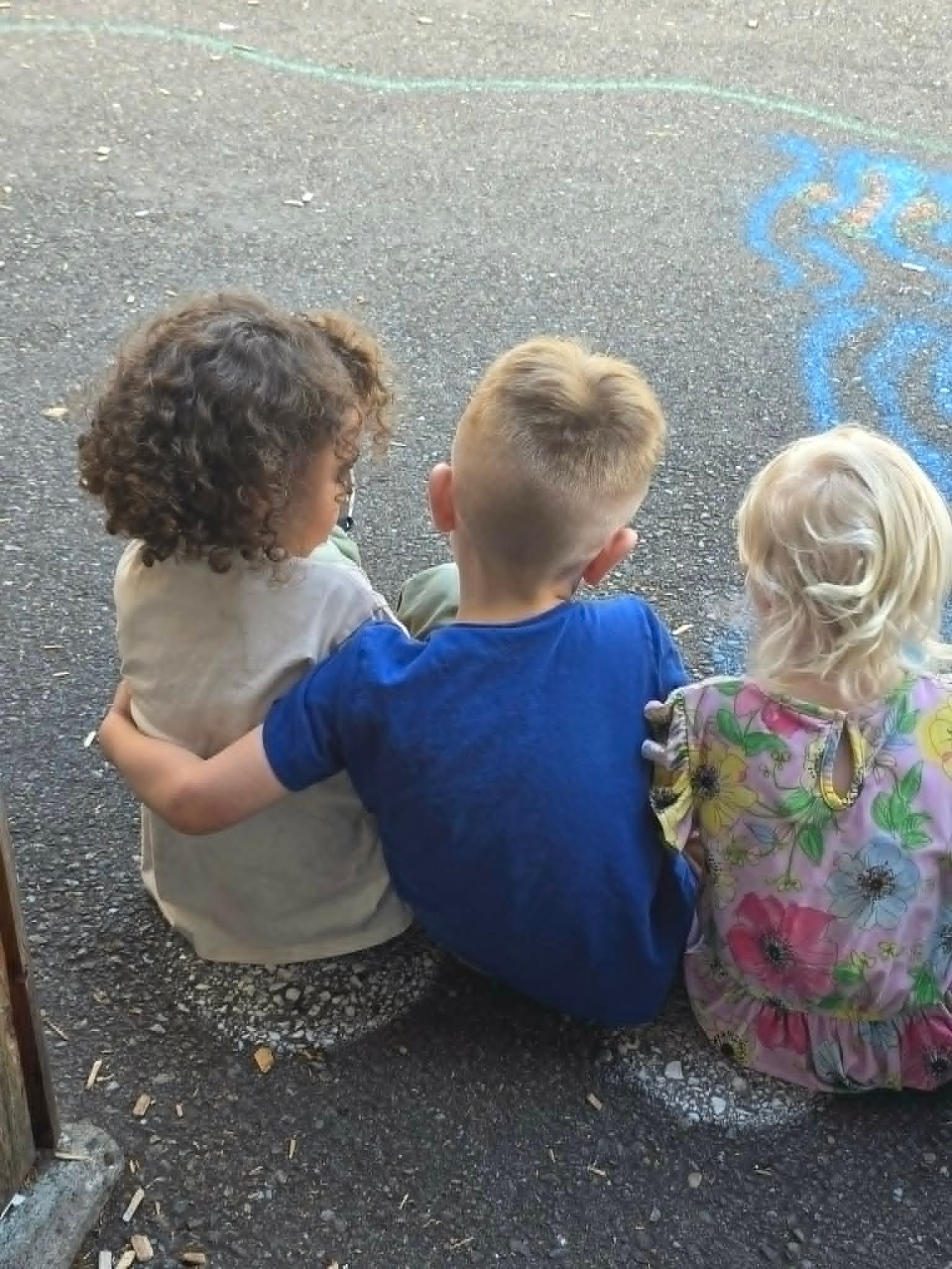 Three children sit closely together on pavement, seen from behind, with chalk markings nearby.