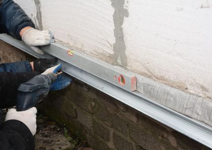 Worker installing a metal flashing strip along the base of an exterior wall with a level tool.