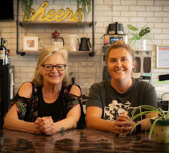 Two women are sitting at a table in front of a sign that says cheers.