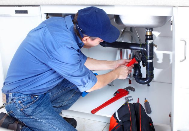 Plumber in blue uniform repairing under-sink pipe with red wrench under a white cabinet