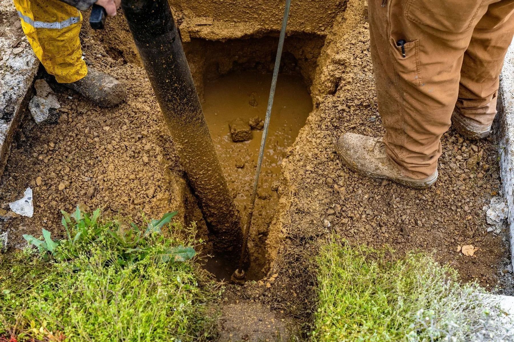 Workers standing around a narrow trench with muddy water and a vertical pipe in a gravel-covered excavation