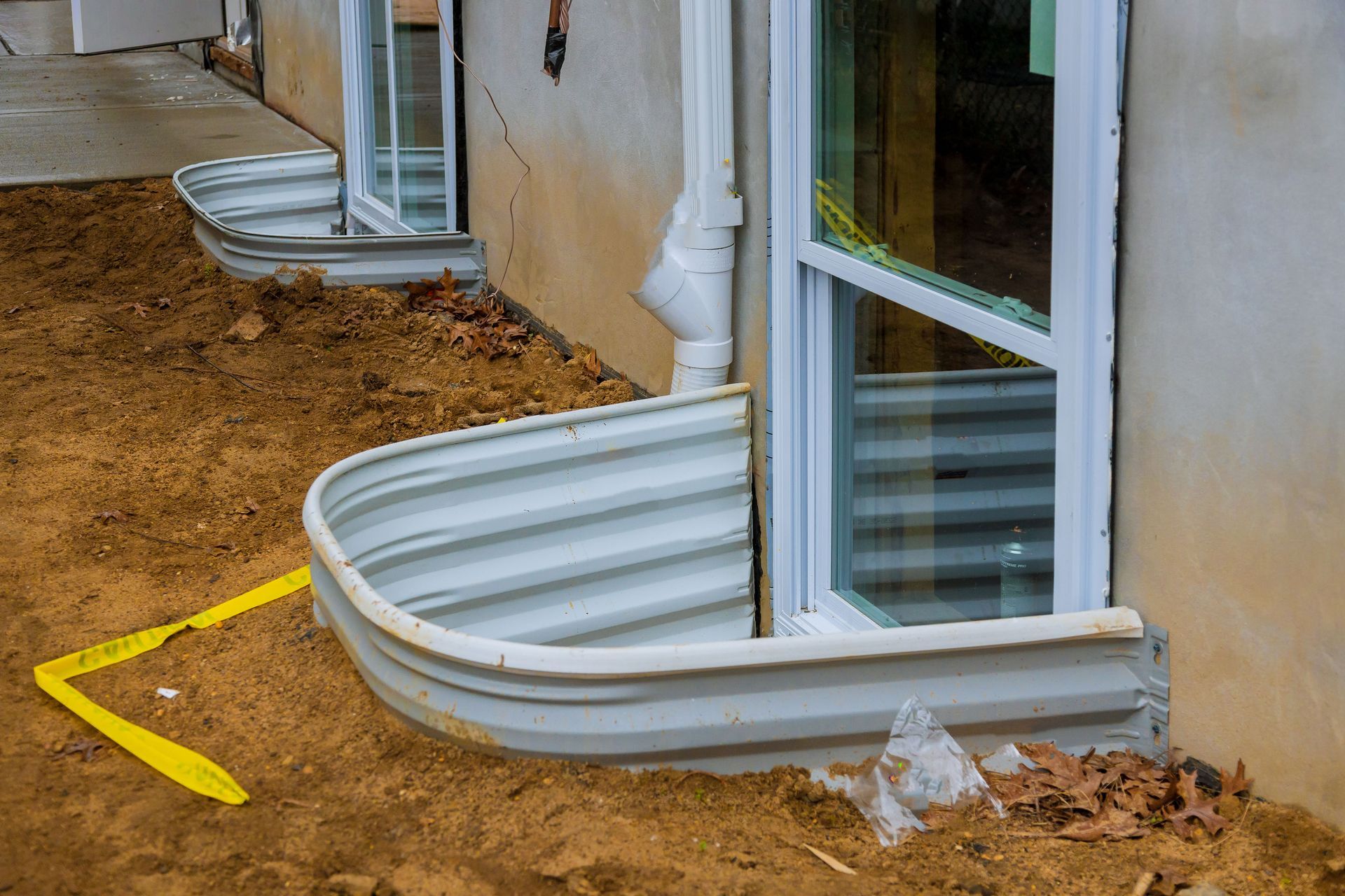 Basement windows with metal window wells, in a home's foundation. The ground is dirt.