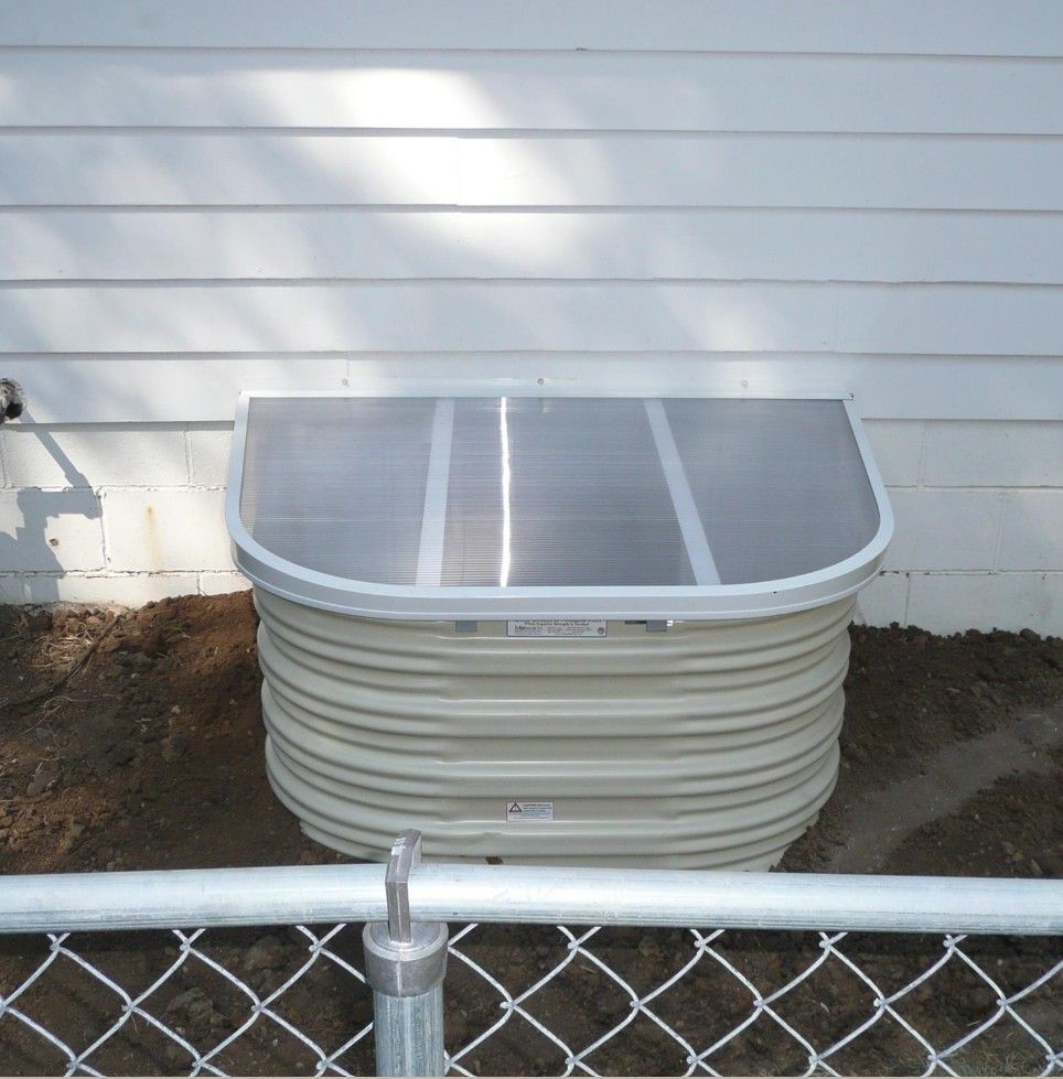 A window well with a clear cover is attached to a white house.