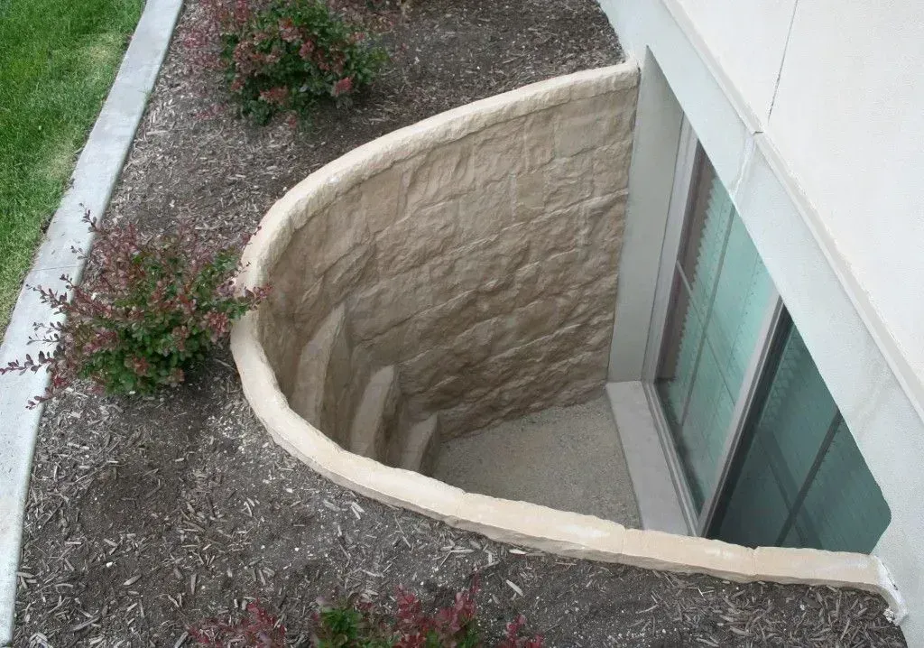 A curved, tan-colored window well with steps, next to a building and landscaped with mulch and plants.