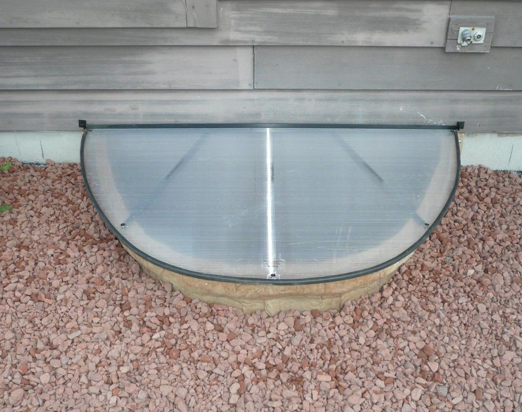 Clear, curved window well cover over a stone-lined window well against a gray wall.