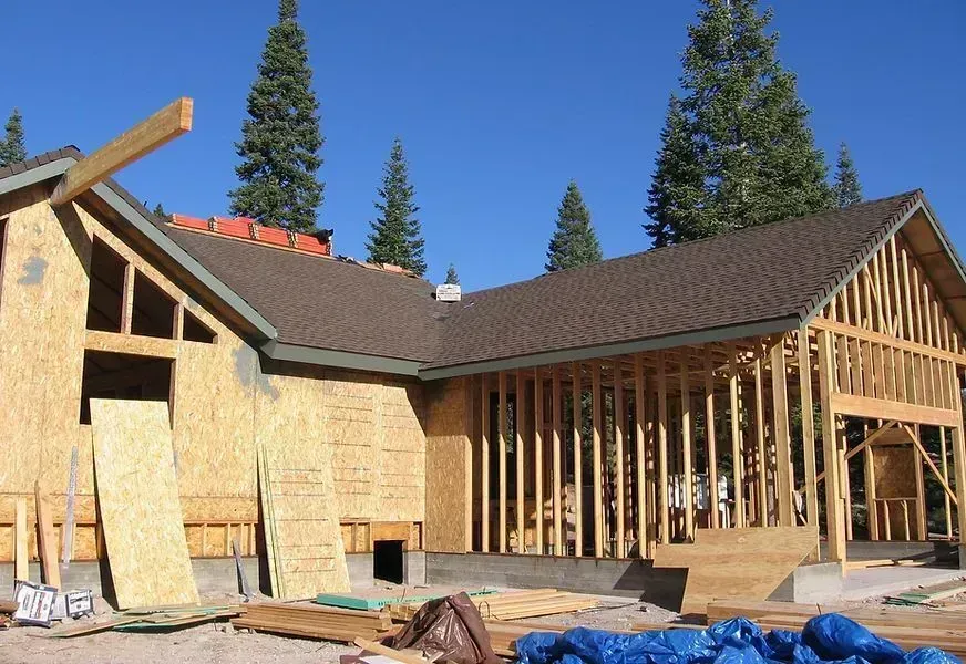 House under construction with exposed wooden framing and dark shingle roof, set against a backdrop of trees and a blue sky.