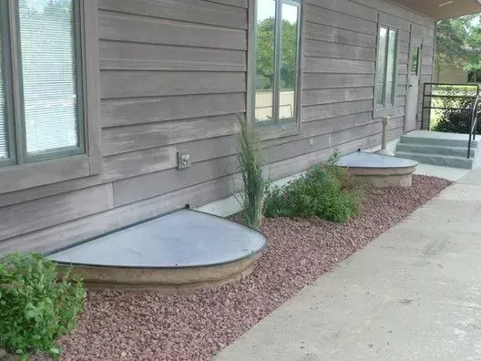 Exterior of a house with window wells covered by gray metal. Brown siding, red gravel.