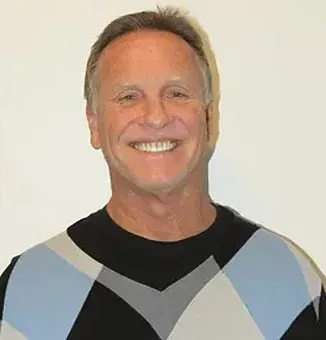 Man smiling, wearing a blue and black argyle sweater, against a white backdrop.
