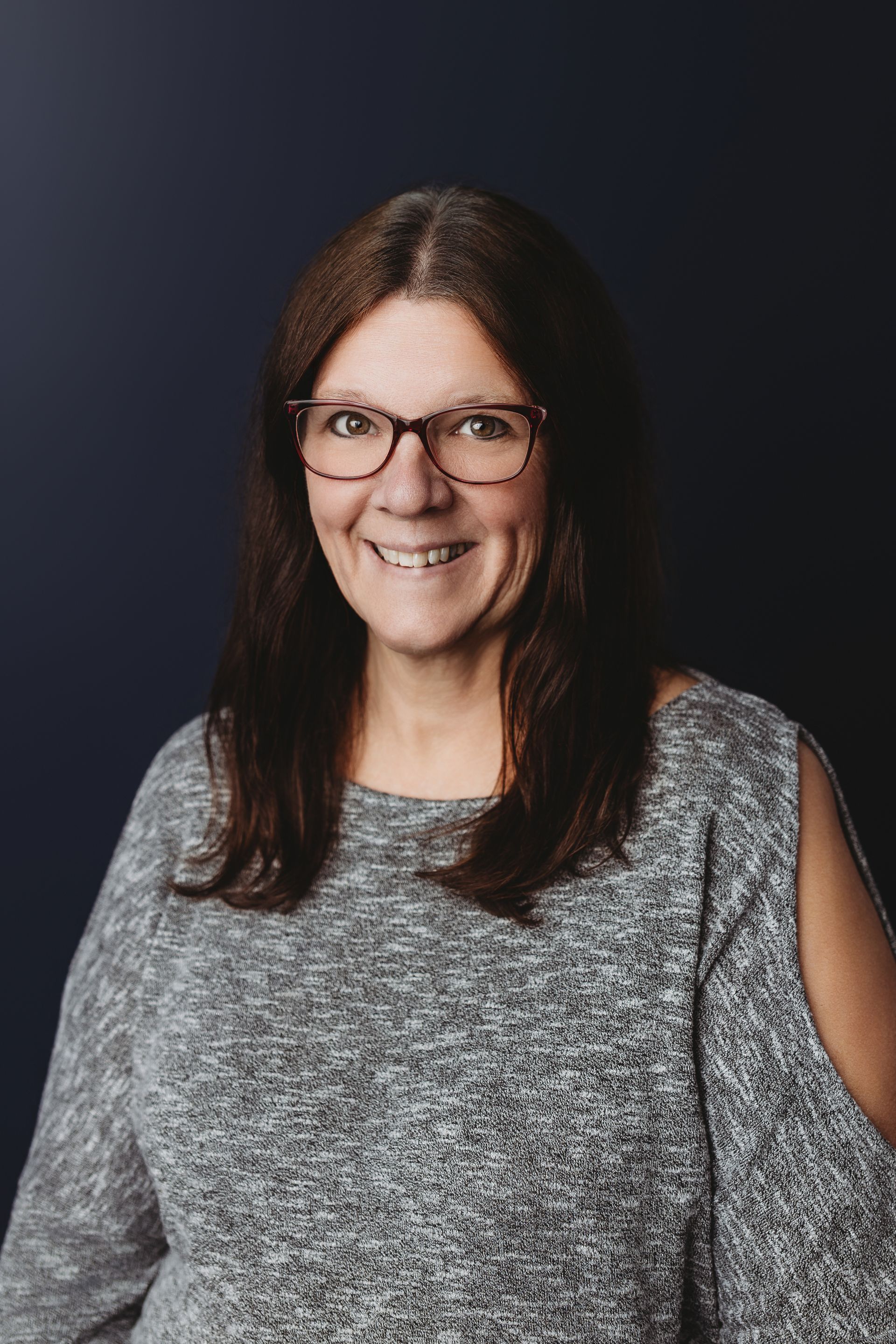Woman with glasses and dark hair smiling in a black sweater against a white wall.