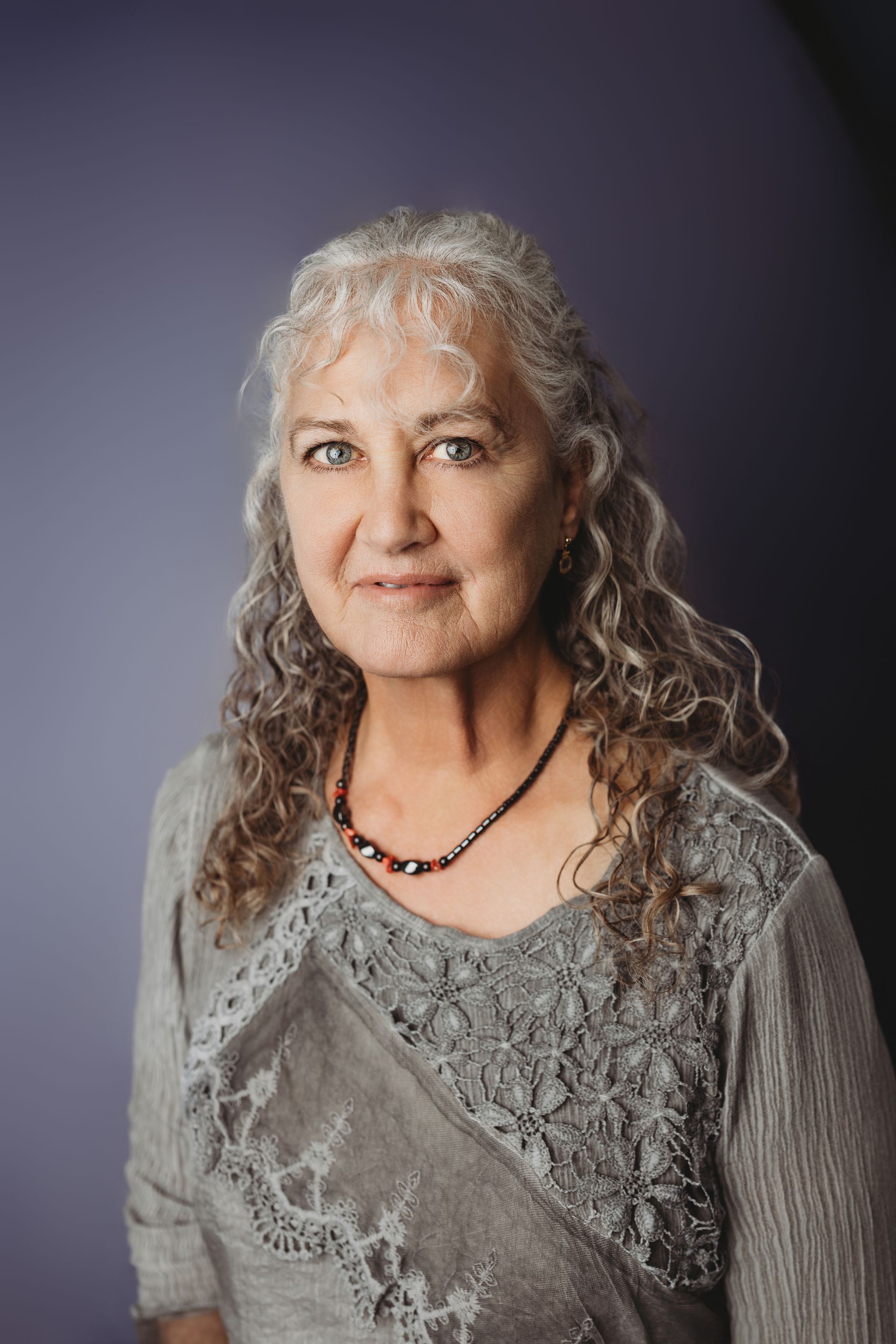 Woman with long, curly gray hair smiles, wearing a black sweater and light gray turtleneck against a white wall.