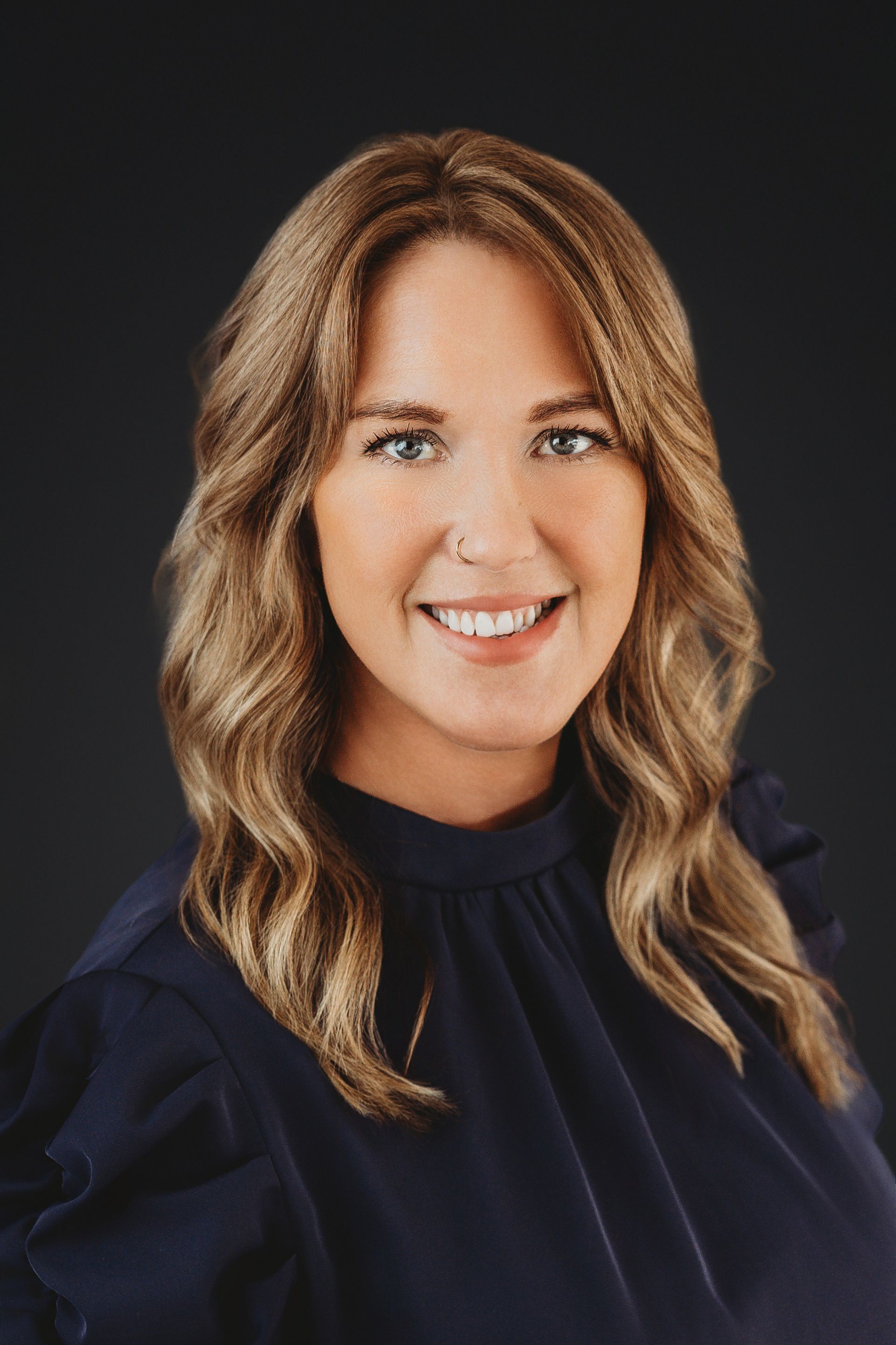 Woman smiling, wearing a black top and sweater, and a necklace. Against a neutral wall.