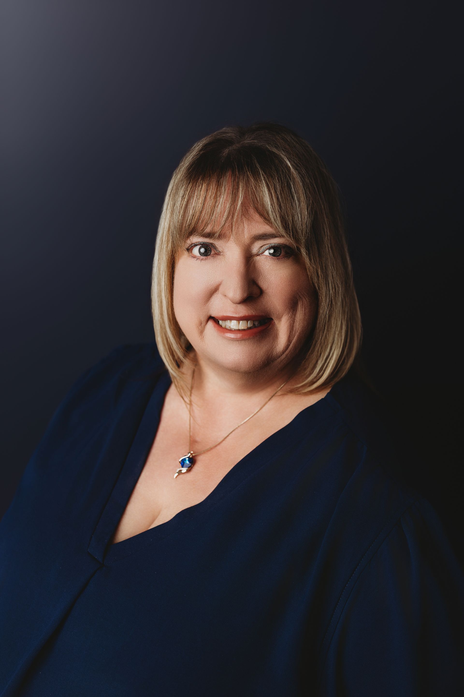 Woman with blonde bangs smiling, wearing a black top with white embroidery, indoors.