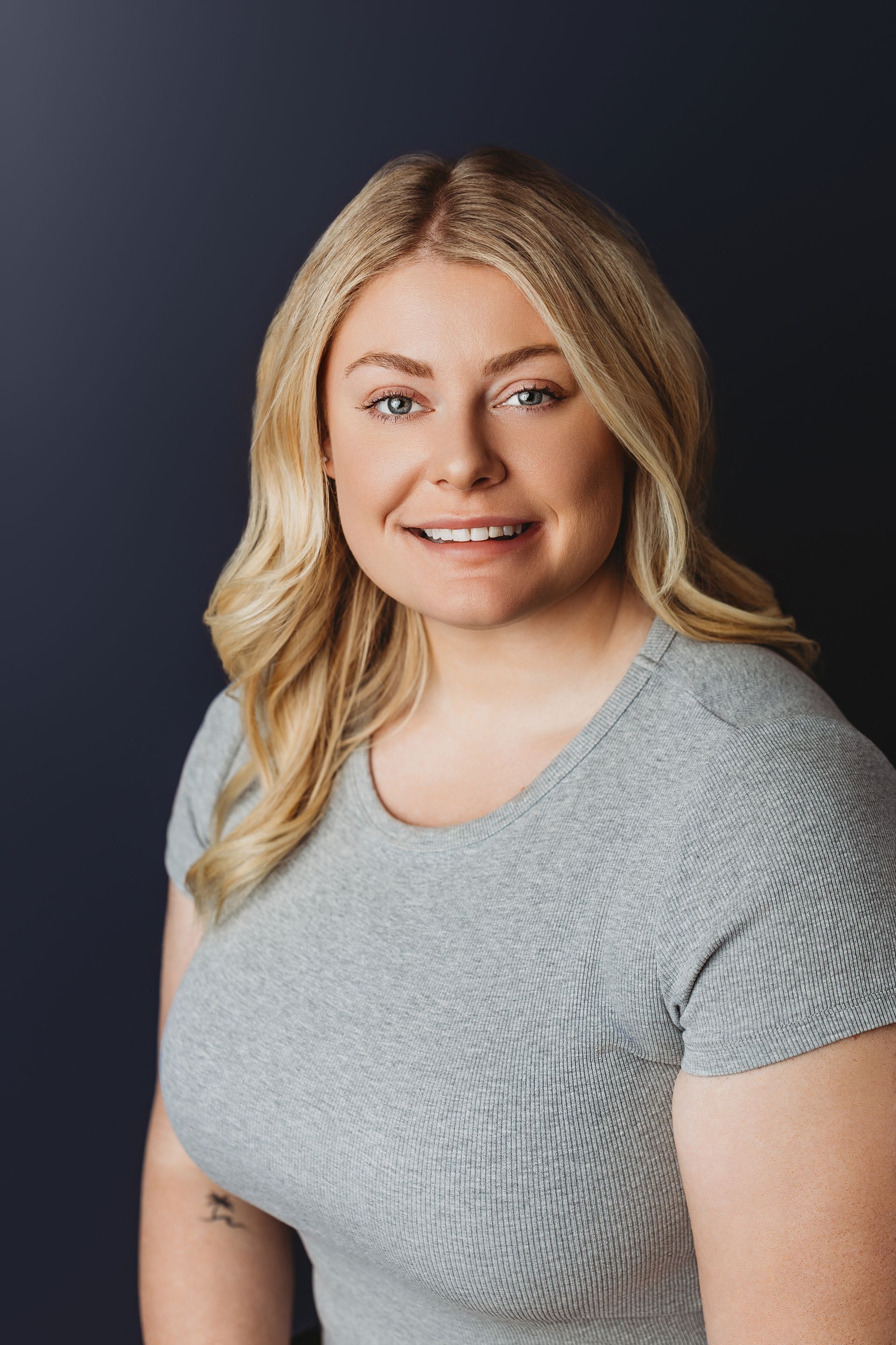 Woman with long blonde hair, smiling, wearing a black blazer and black top in front of a white textured wall.