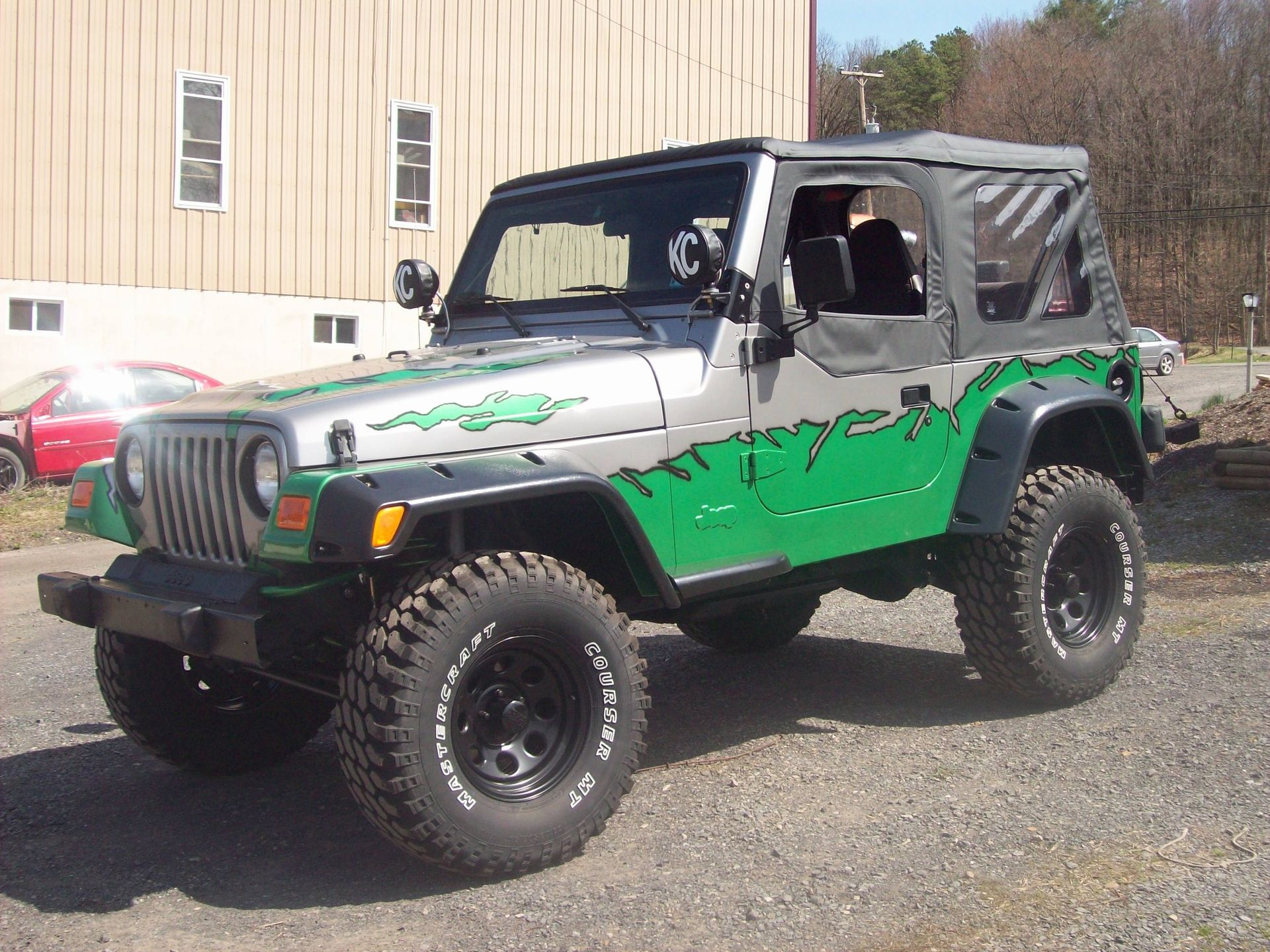 Silver and green Jeep Wrangler with black wheels, soft top, parked in front of a tan building.