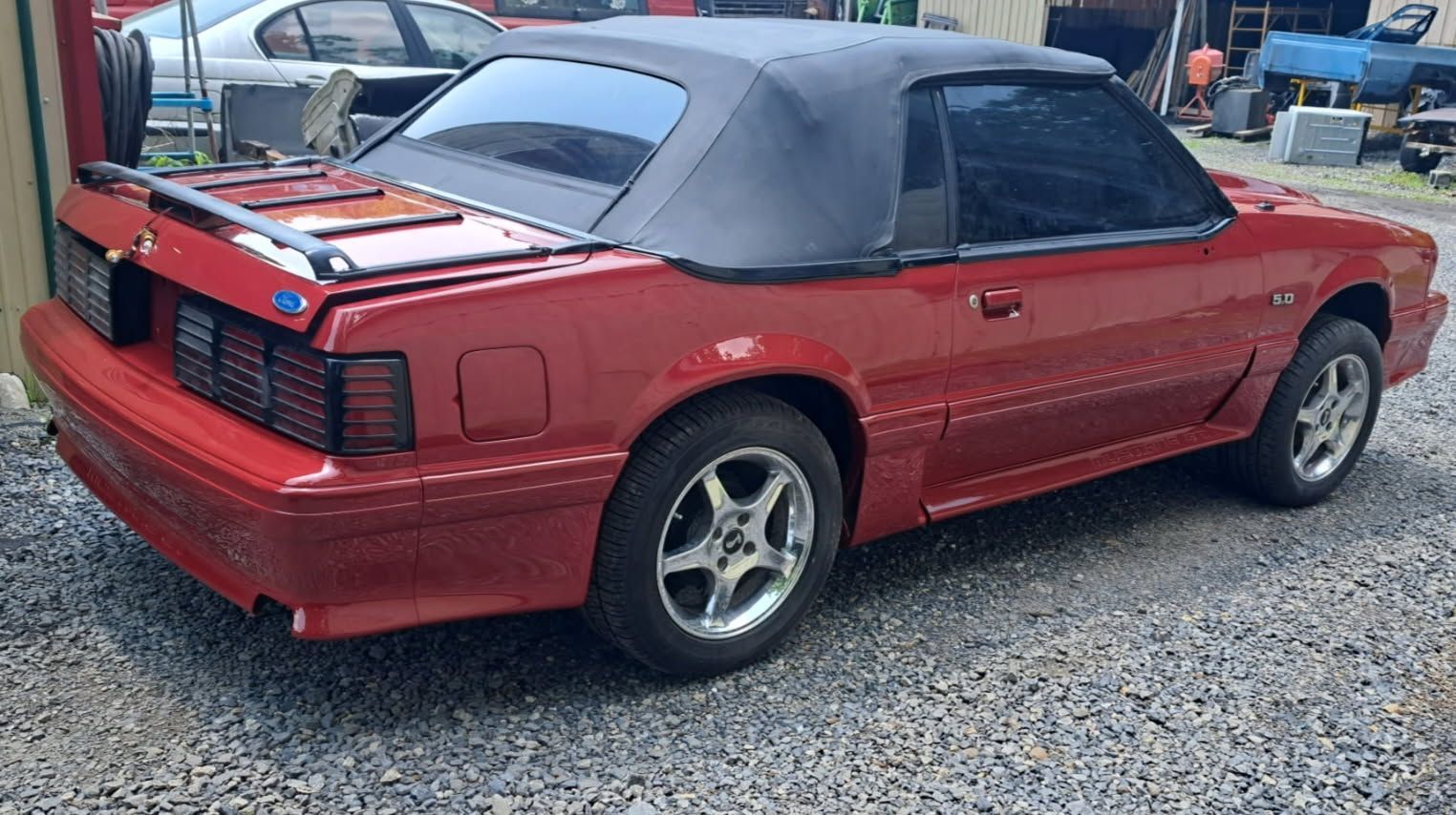A red Ford Mustang convertible with a black soft top parked on a gravel surface.