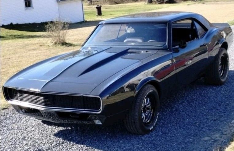 Black 1967 Chevrolet Camaro muscle car with a dark hood scoop, parked on gravel.