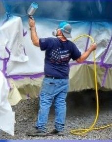 Person in blue shirt and jeans sprays paint on a vehicle, wearing a respirator.