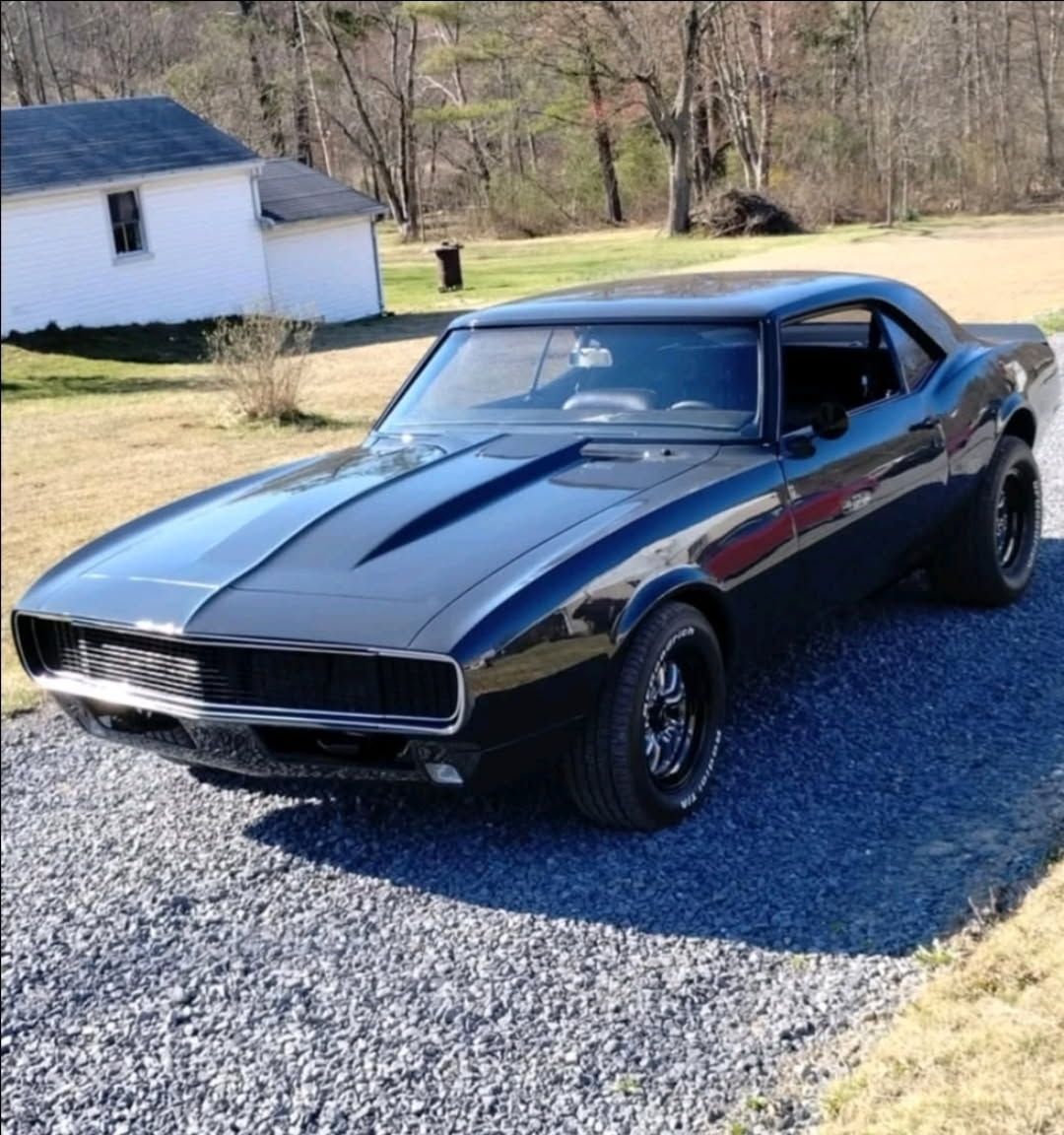A glossy black 1967 Chevrolet Camaro parked on a gravel driveway in front of a white building and bare trees.